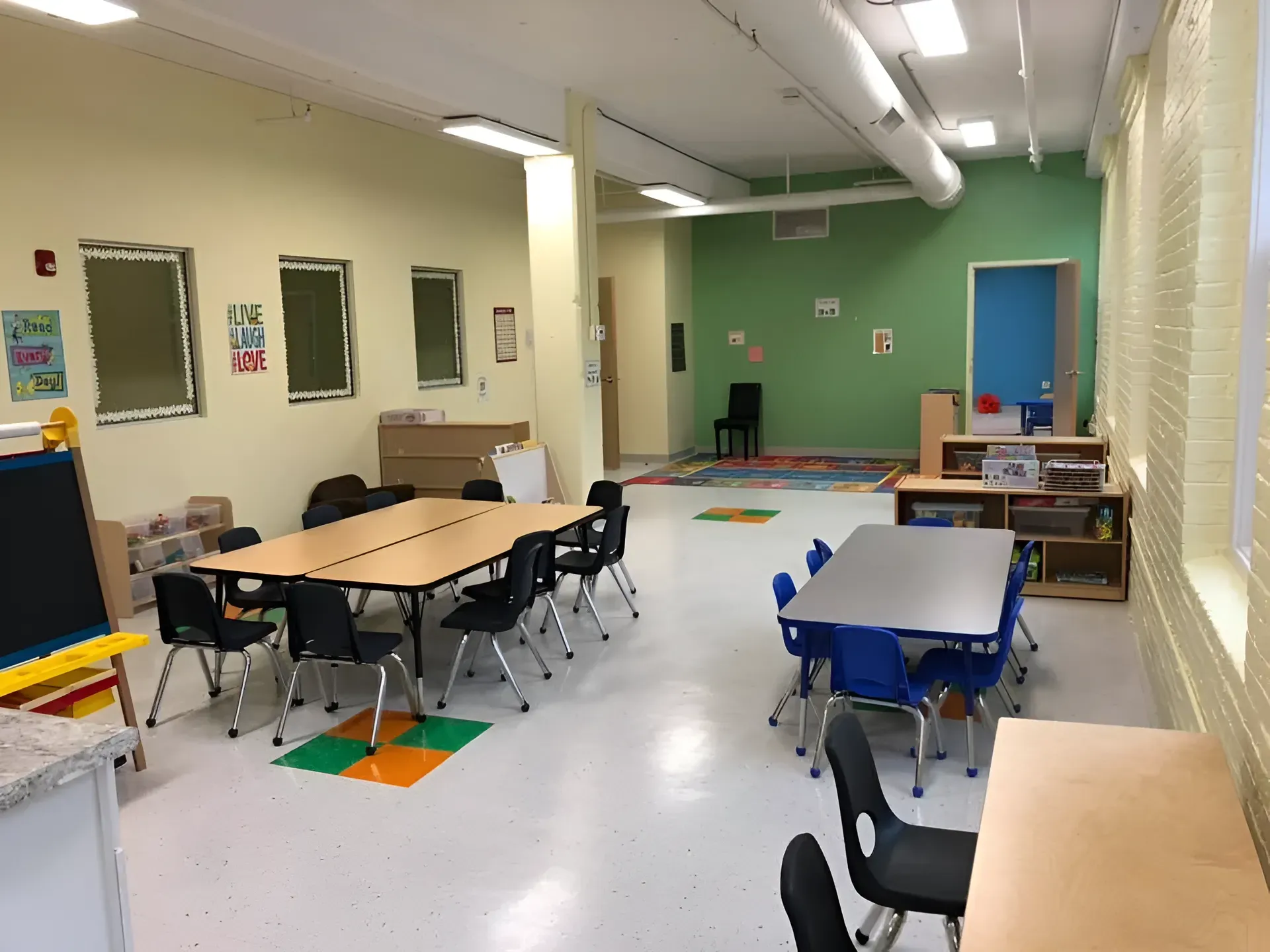 Empty classroom with tables, chairs, and green walls under fluorescent lights.