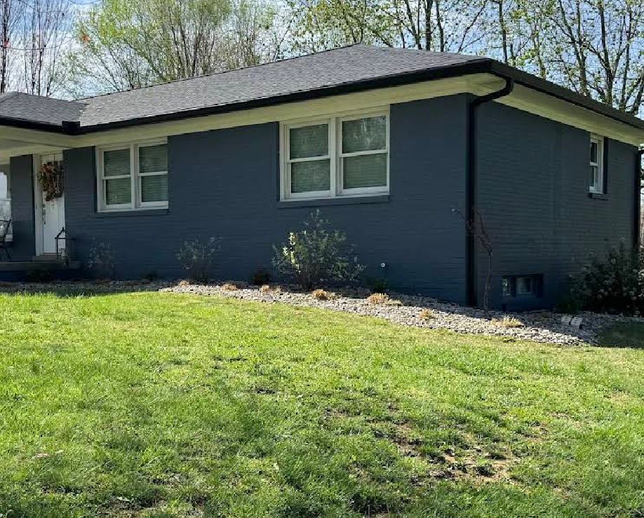 Blue-painted brick house with white trim and a dark roof; front yard with green grass and landscaping.