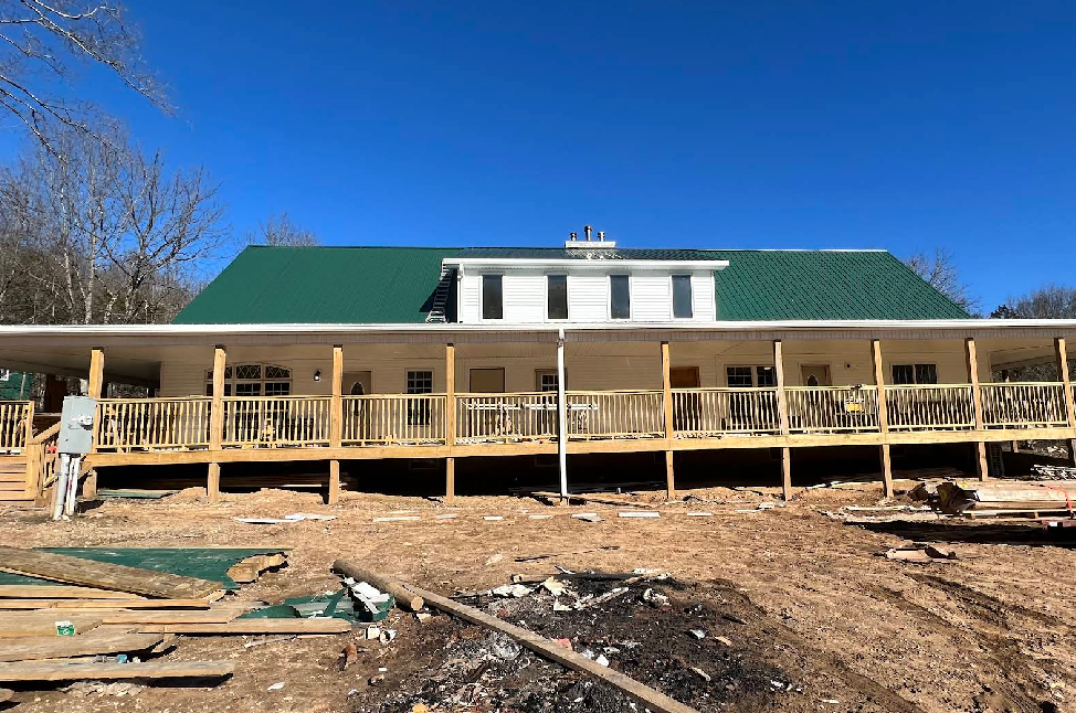 A building with a green roof, wrap-around porch, and wooden deck under construction against a clear blue sky.