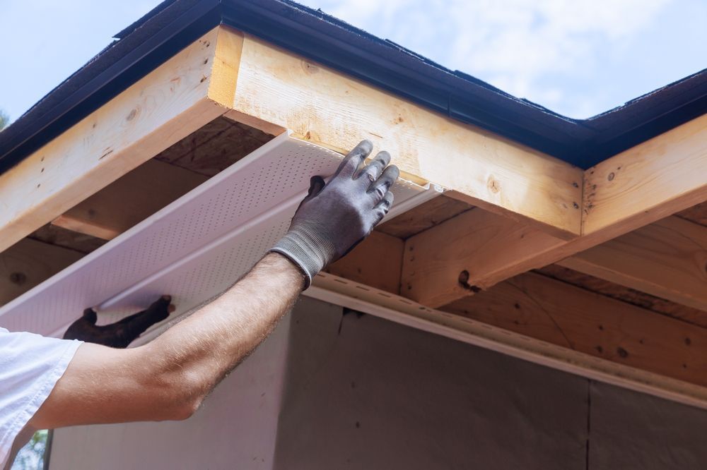 Person in work gloves installing white perforated soffit panel on wooden structure.