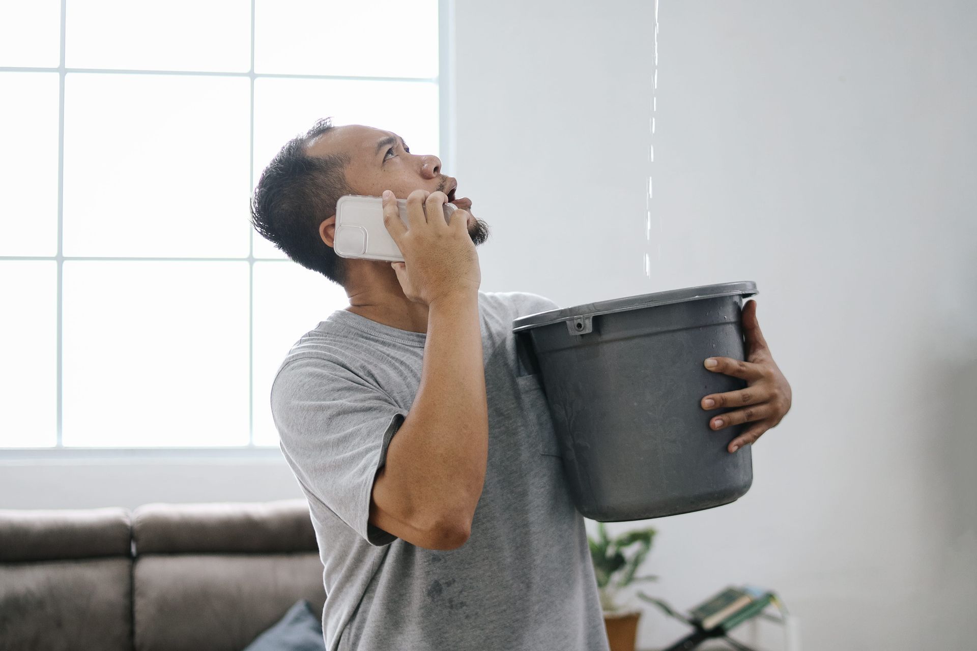 Man on phone holding a bucket, looking up as if reacting to a leak indoors