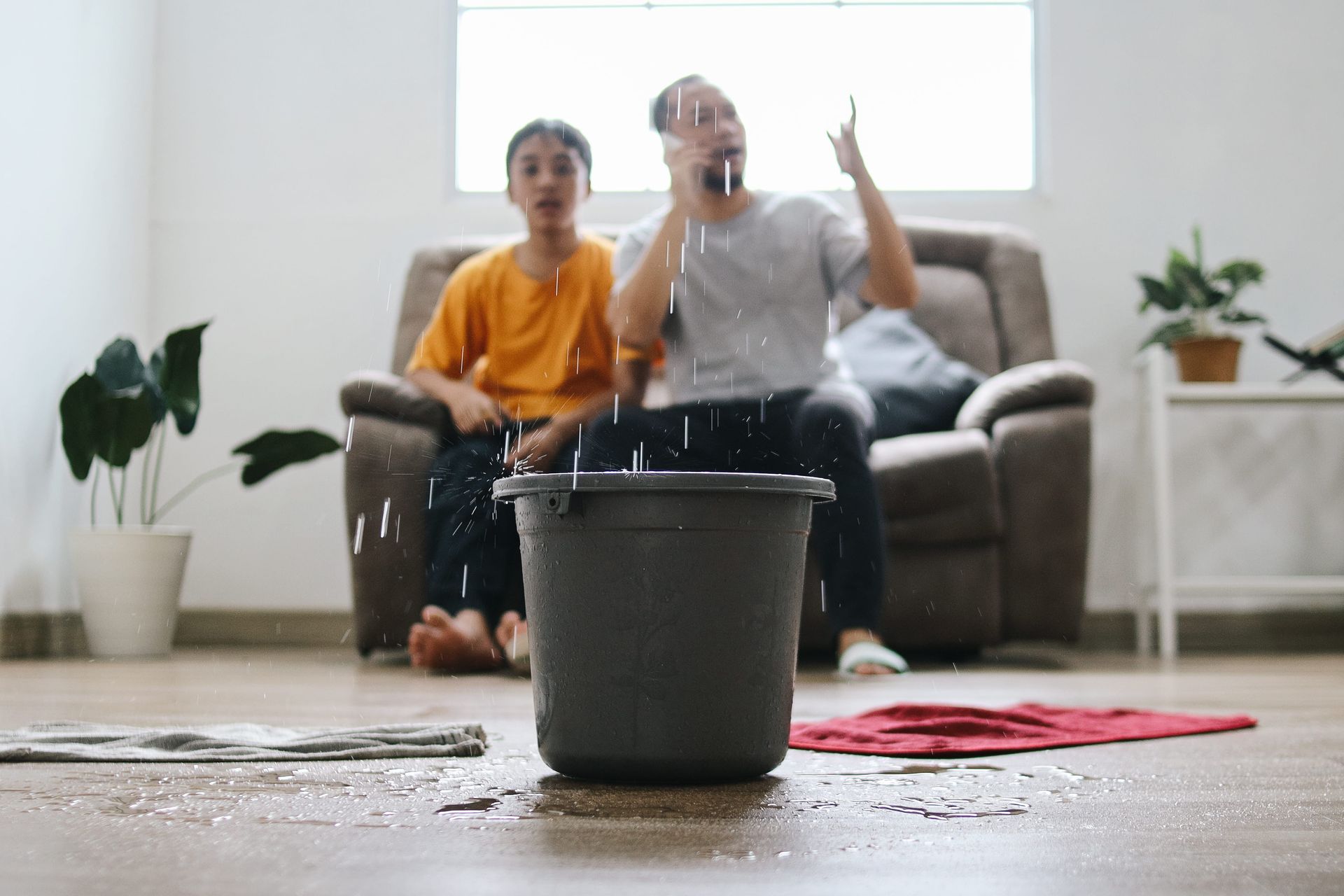 Two people on a couch in a messy living room as water splashes from a bucket in the foreground