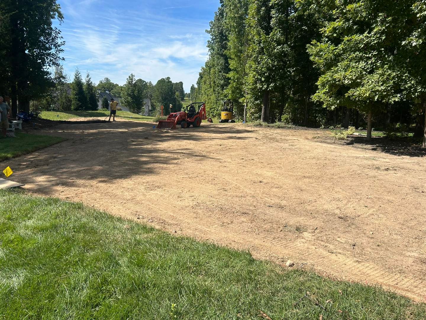 Dirt clearing in a wooded park with a red tractor, workers, and green grass in the foreground