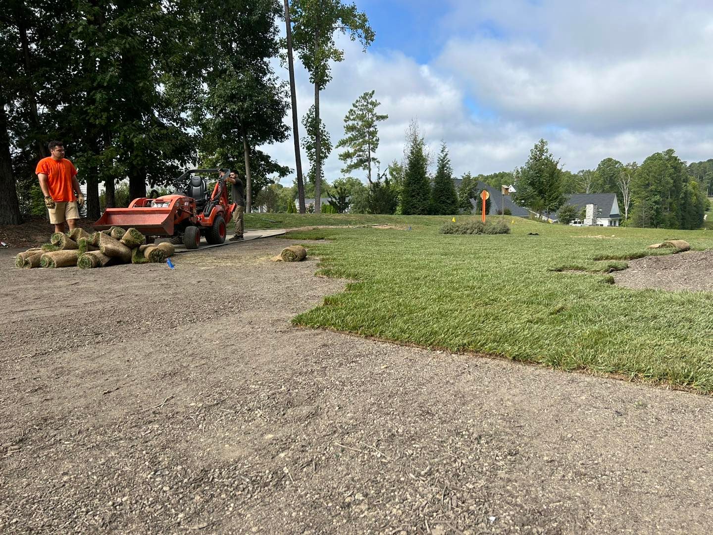Two workers with an orange tractor spreading gravel in a grassy park under a cloudy sky