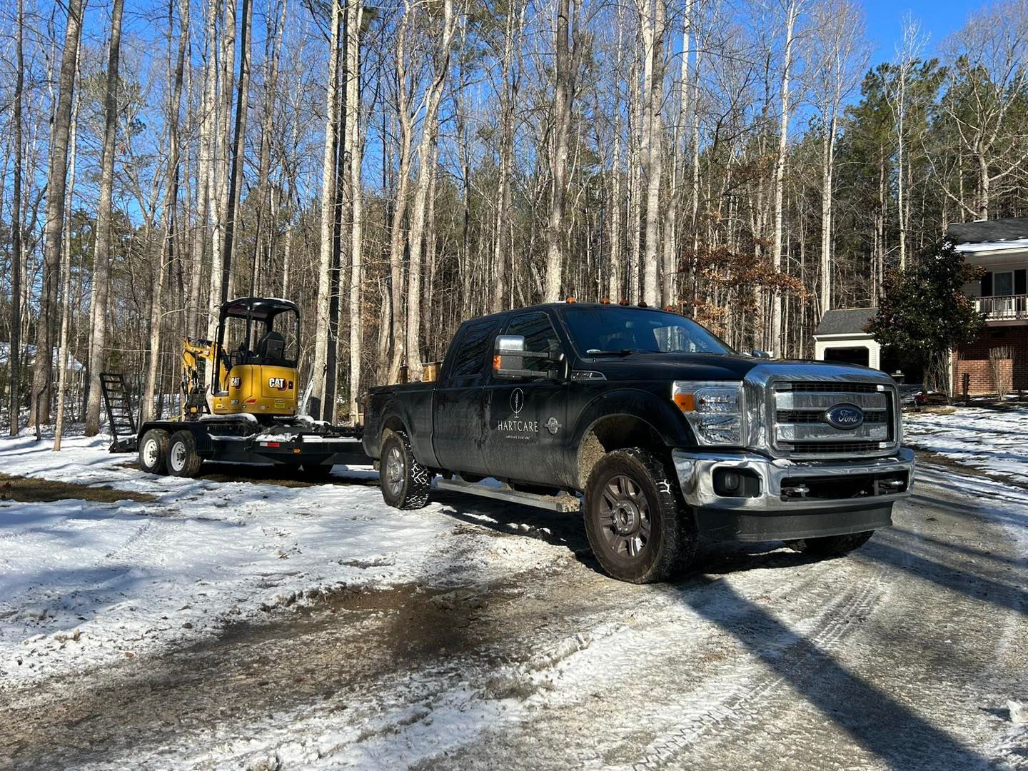 Black pickup truck towing a yellow mini excavator on a snowy driveway in a wooded area