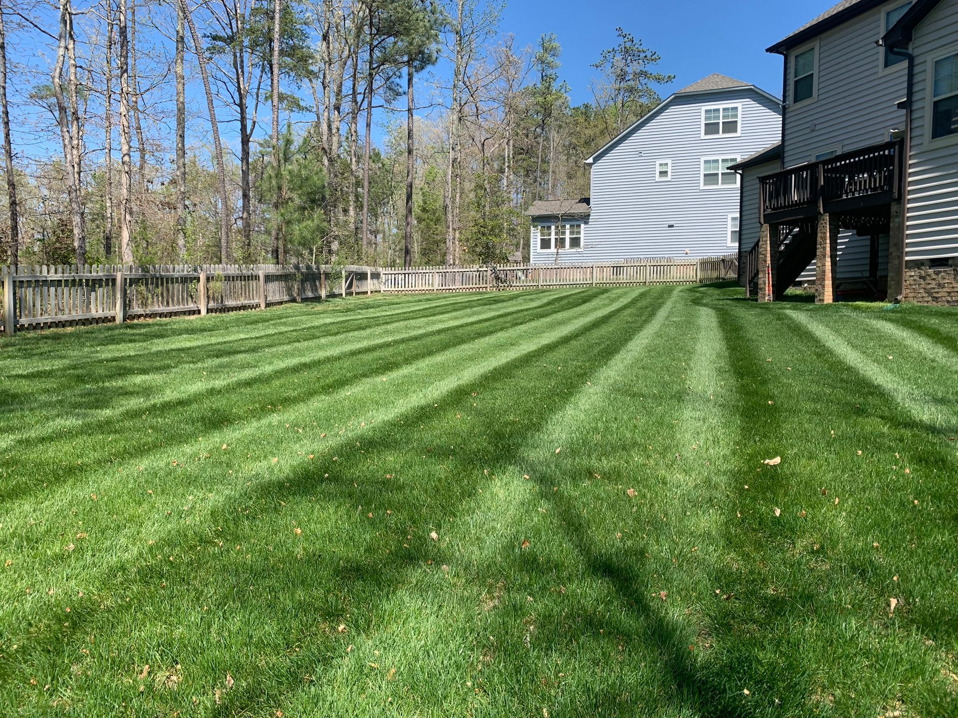 A well-manicured lawn with distinct, parallel mowing stripes, extending toward a house with a wooden deck and woods beyond.