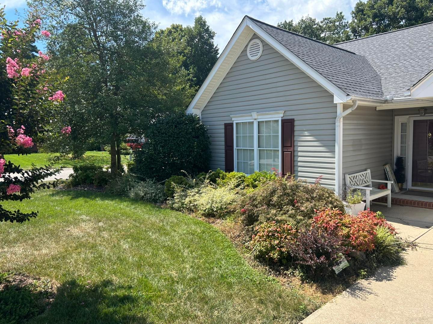 Suburban house exterior with beige siding, front lawn, shrubs, and a flowered garden in bright sunlight