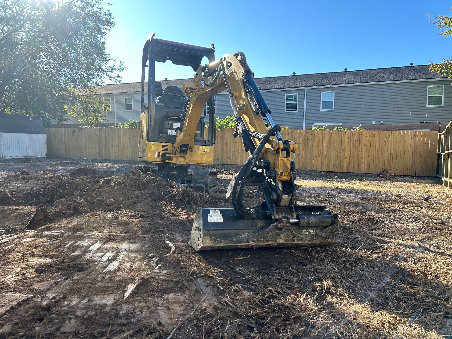 A yellow Caterpillar mini excavator with a mulching attachment parked on dirt in a fenced residential lot.