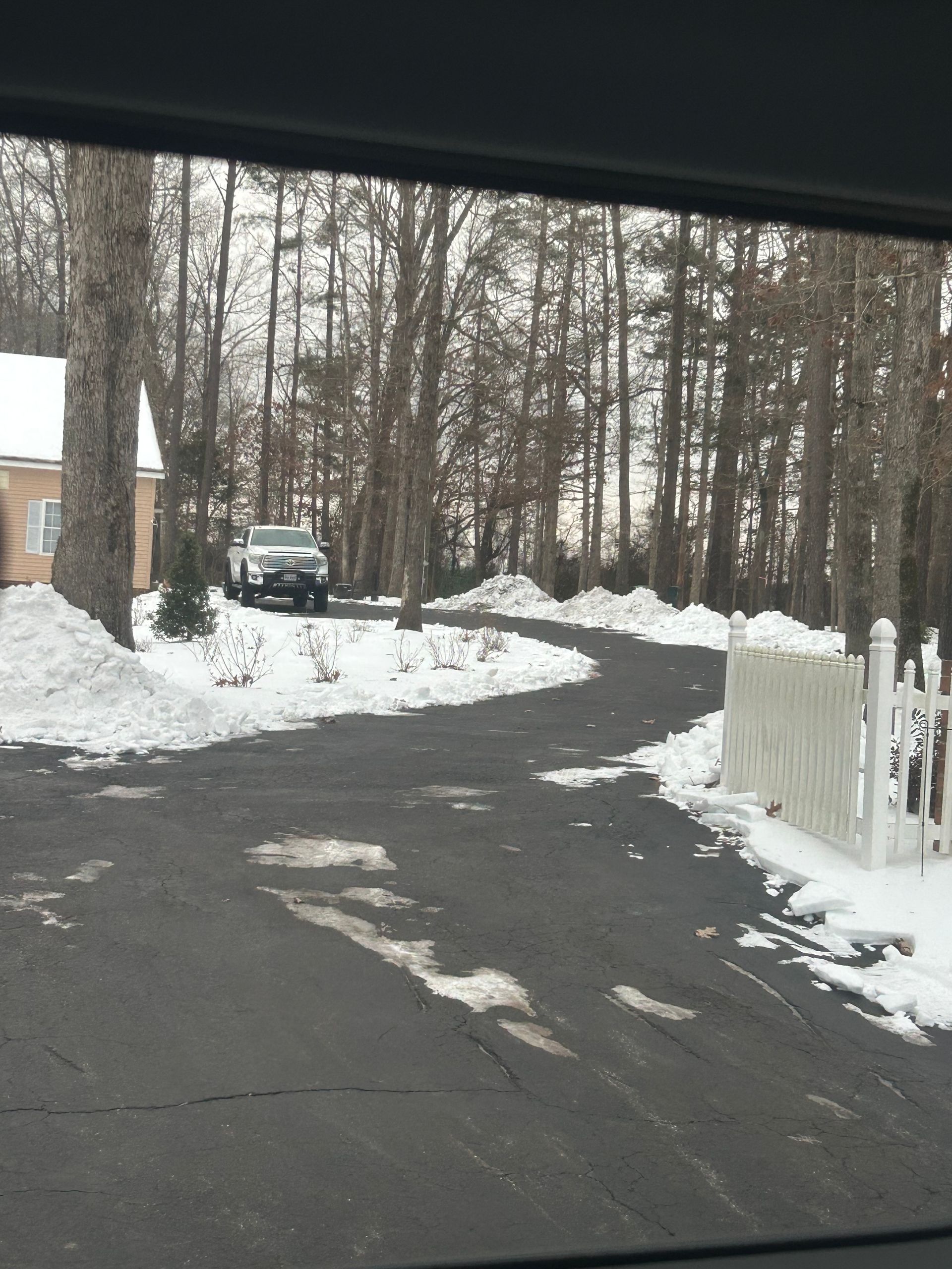 A snowy driveway leads past a white fence toward a house and a car among bare trees on a cloudy winter day.