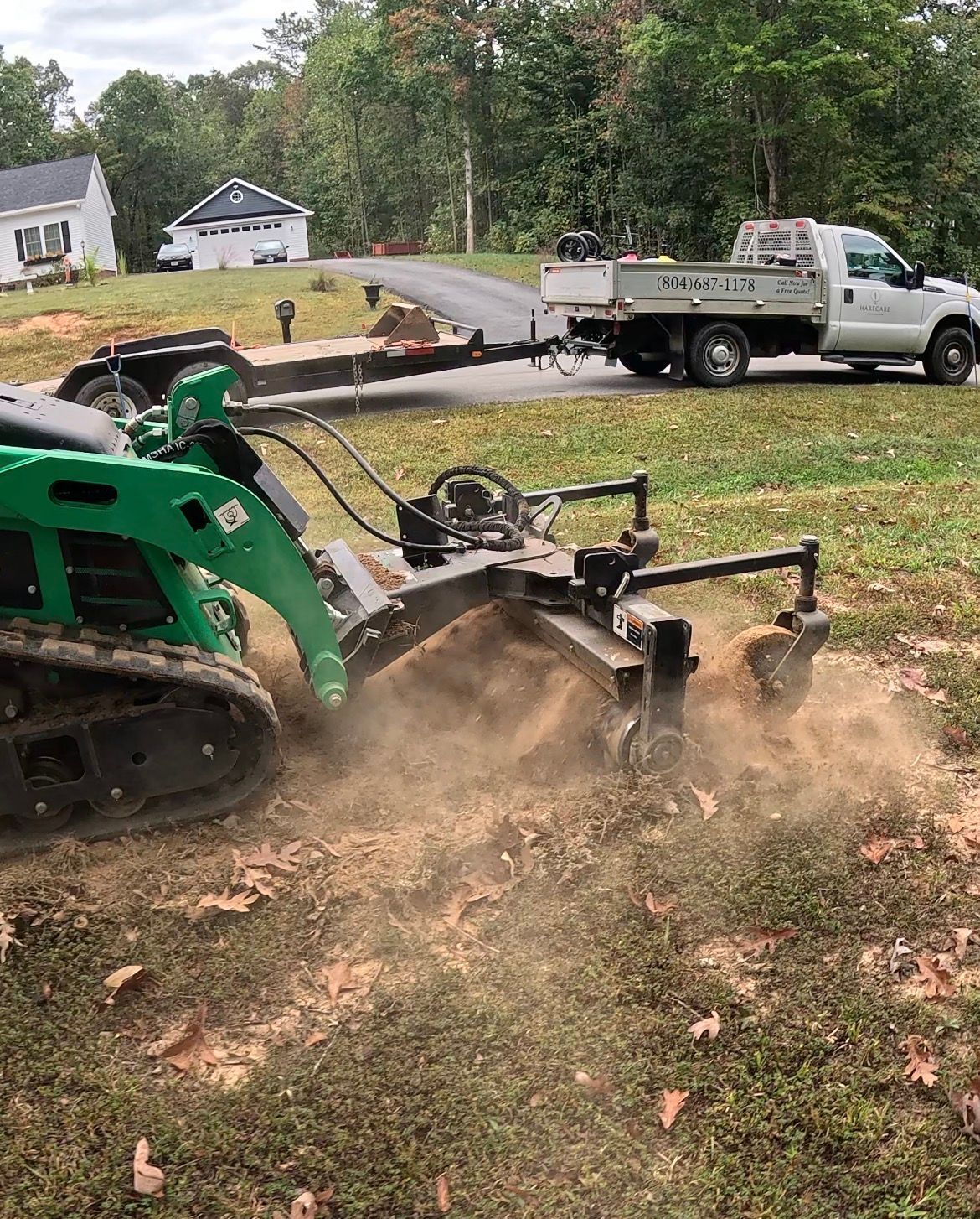Green skid-steer with attachment digging dirt beside a white utility truck in a yard, dust rising.