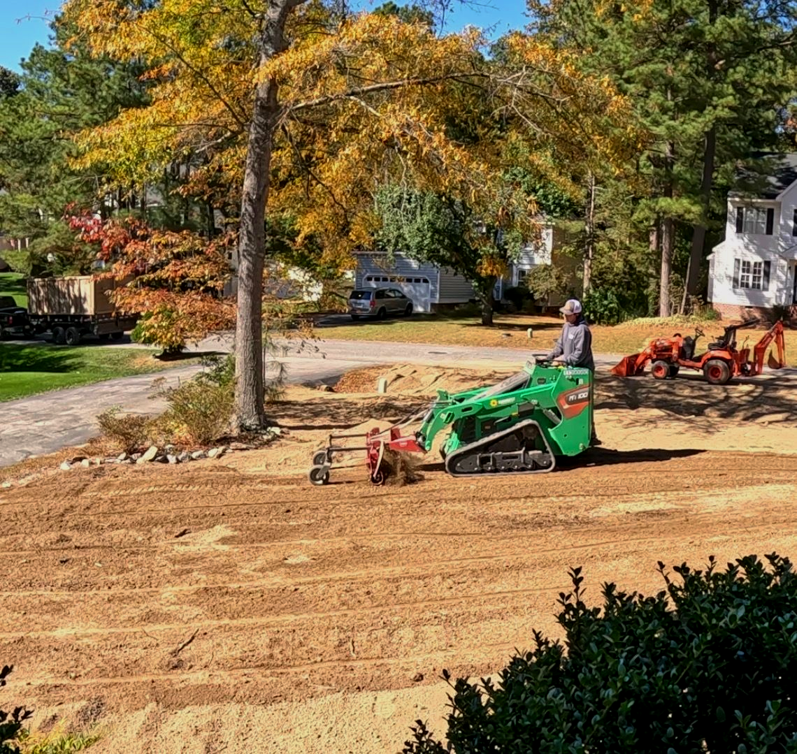 A person operating a yellow Caterpillar mini excavator to dig up shrubs in a residential front yard.