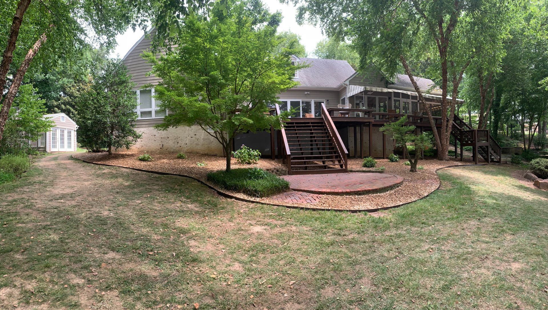 A multi-level house with a wooden deck and staircase, featuring a stone patio and landscaping in a wooded backyard.