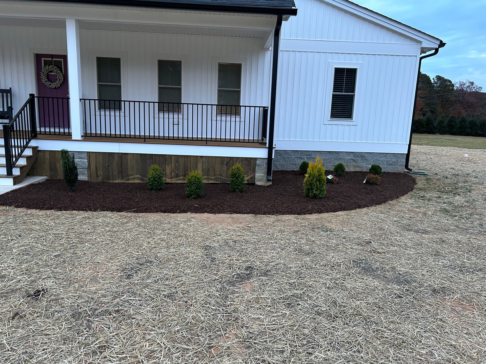 A white house with a covered porch and railing, featuring a freshly mulched garden bed with small evergreen shrubs.