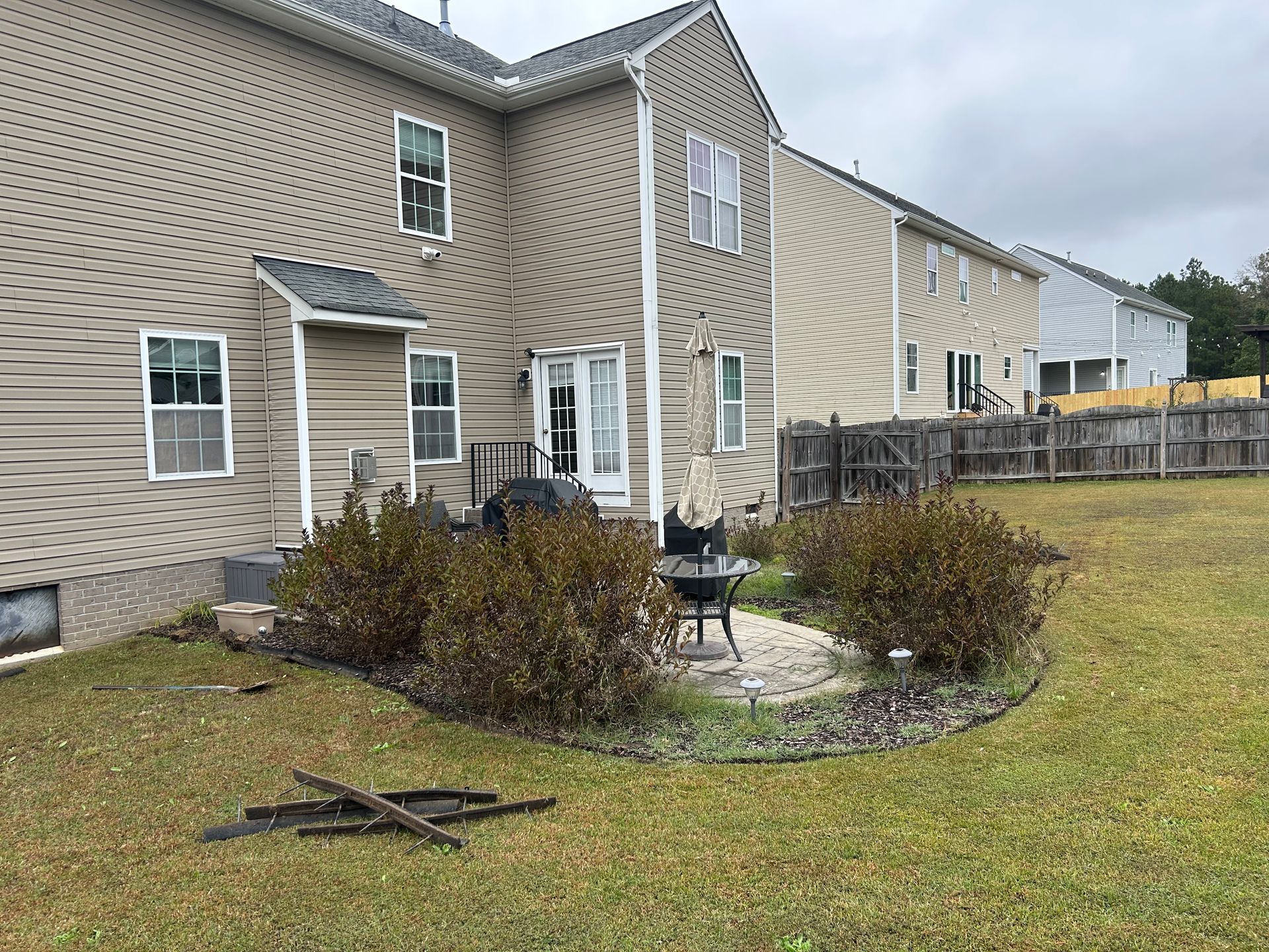 Backyard with a beige townhouse, small patio, shrubs, and a lawn under cloudy skies