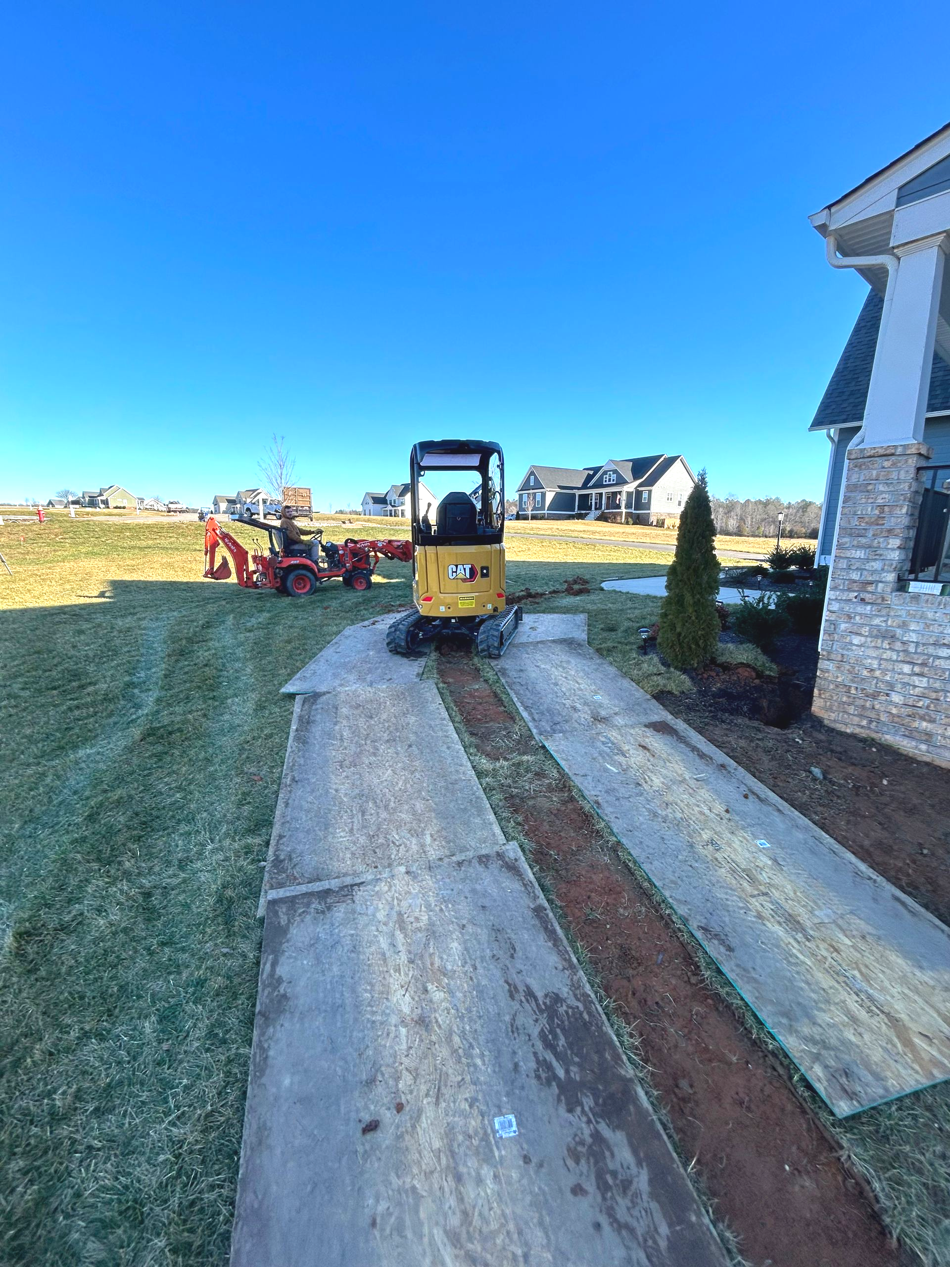 Yellow mini excavator on a gravel driveway beside a house, with a tractor in the yard under a clear blue sky