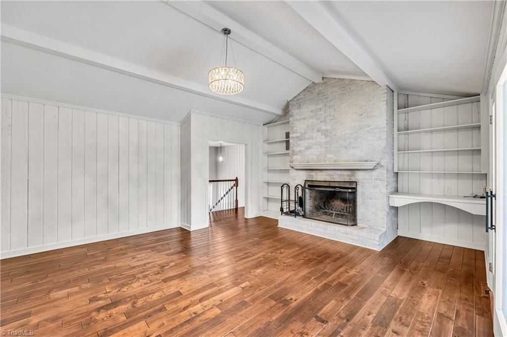 Bright empty living room with hardwood floors, white paneled walls, vaulted ceiling, and a stone fireplace.