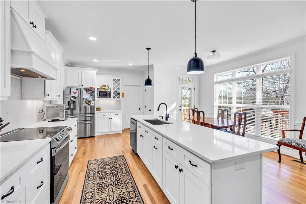 Bright modern white kitchen with large island, wood floors, pendant lights, and stainless-steel appliances.