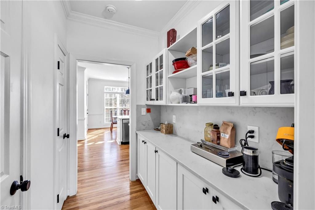 Bright white kitchen with glass cabinets, coffee maker, and a hallway with wood floors leading to another room