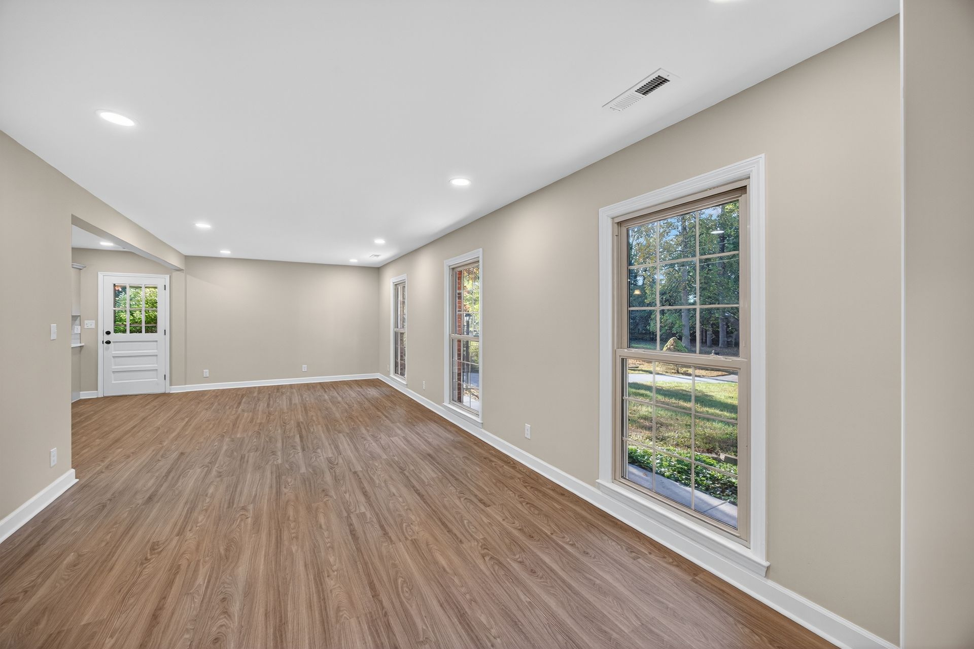 Empty bright living room with wood floors, recessed lights, and large windows looking out to greenery
