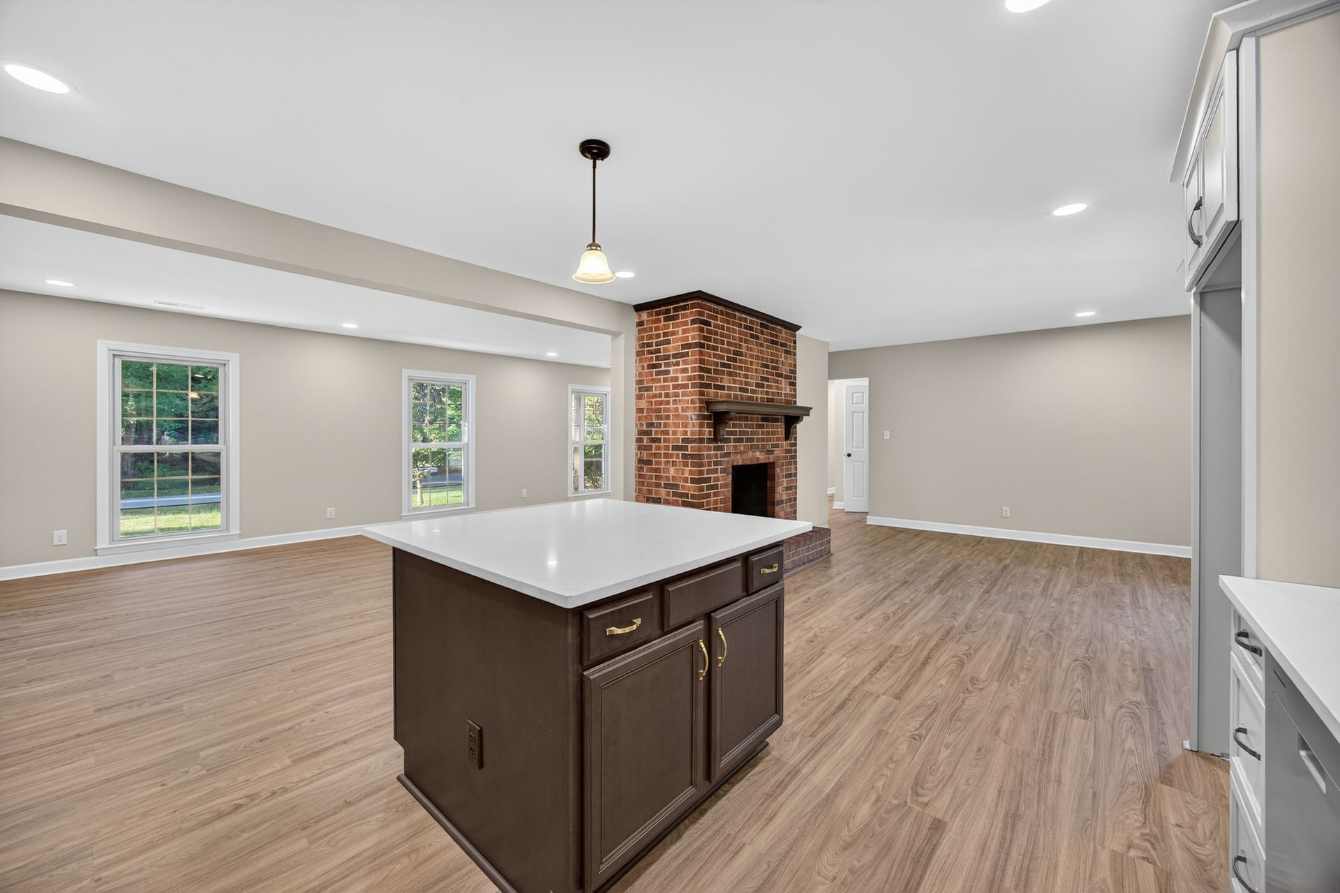 Empty open-plan living area with white walls, wood floors, central island, and brick fireplace