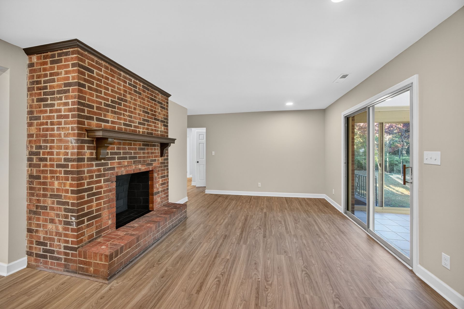 Empty living room with brick fireplace, wood floors, beige walls, and sliding glass doors.