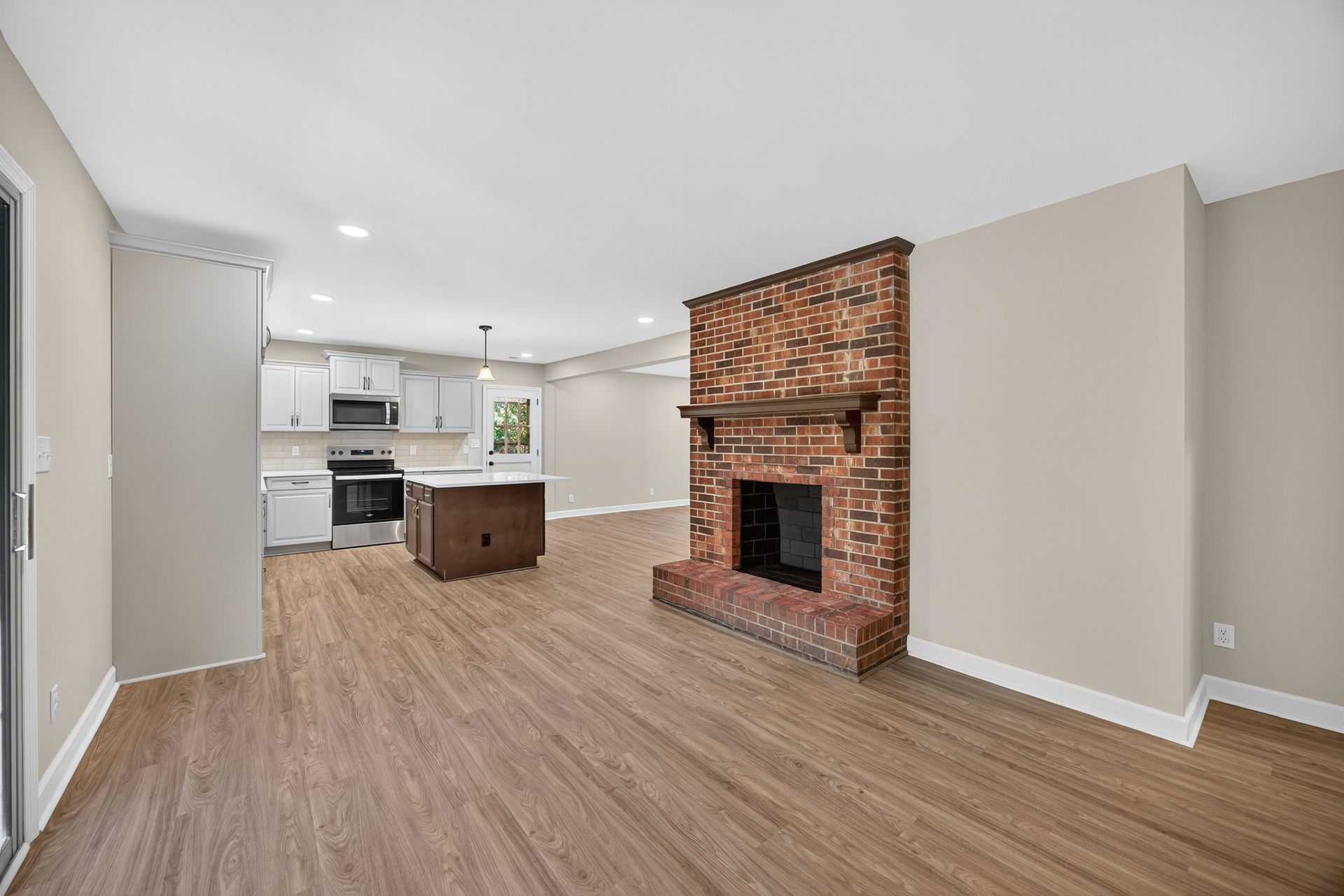 Empty open-plan living room with brick fireplace and kitchen island, hardwood floors, beige walls
