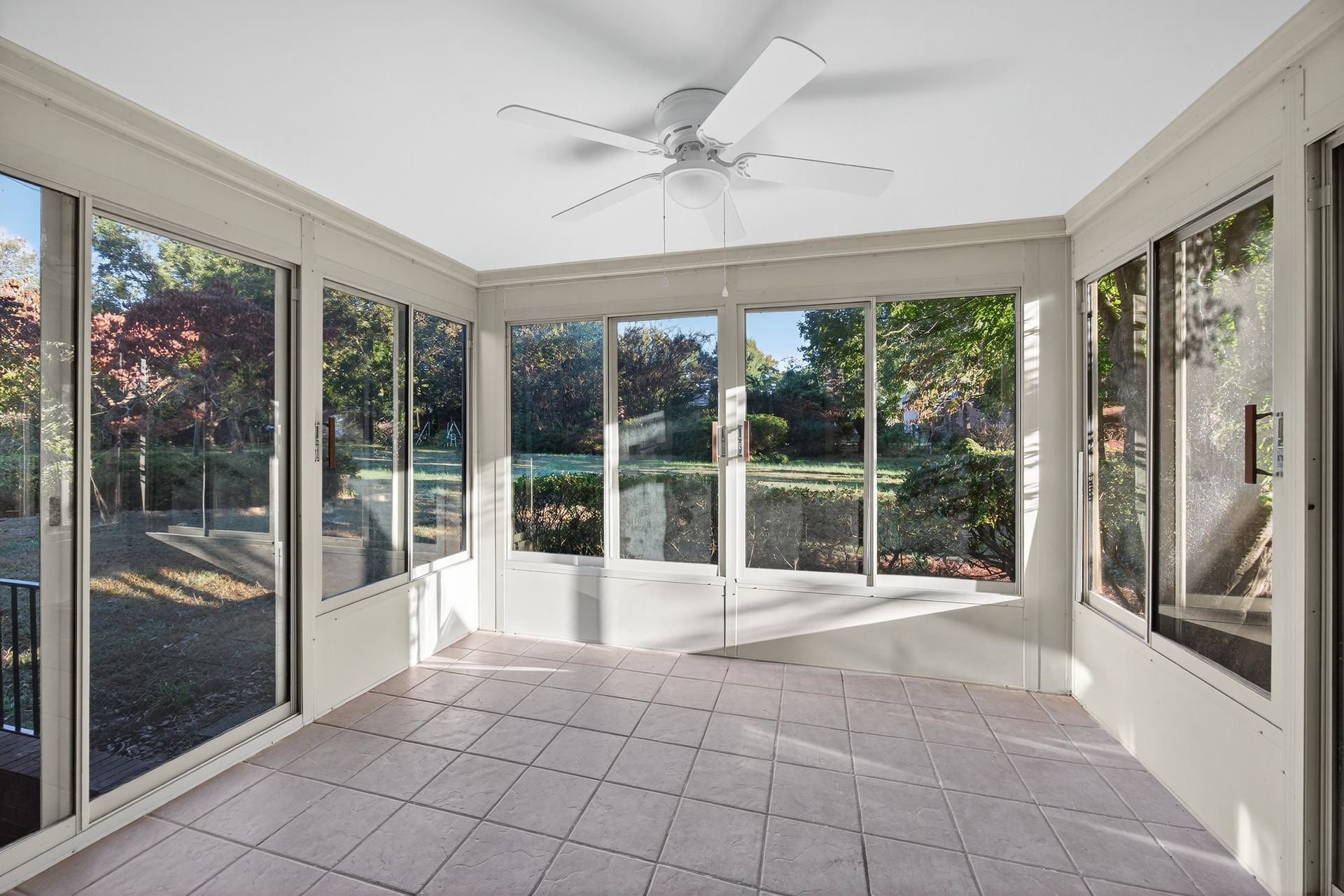 Bright sunroom with tiled floor, large windows, and a ceiling fan overlooking a garden.