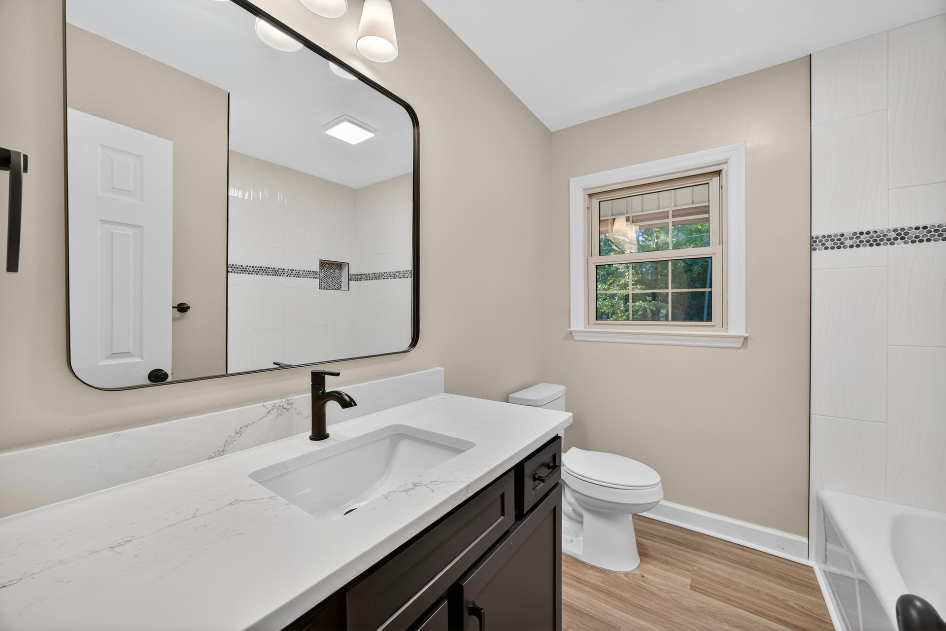 Bright modern bathroom with white vanity, black faucet, mirror, toilet, tub, and small window