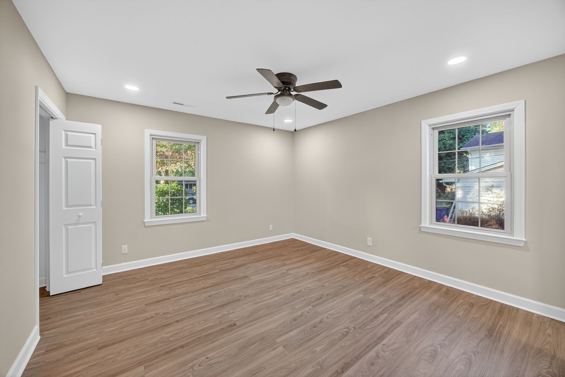 Empty beige bedroom with wood flooring, white trim, two windows, and a ceiling fan.