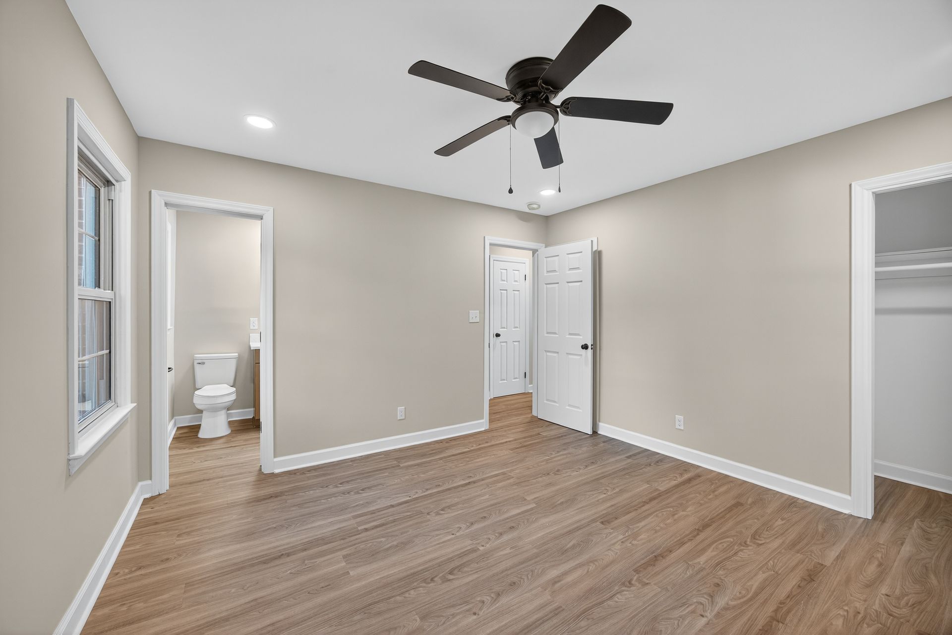 Empty beige bedroom with wood floors, ceiling fan, windows, and an attached bathroom visible.