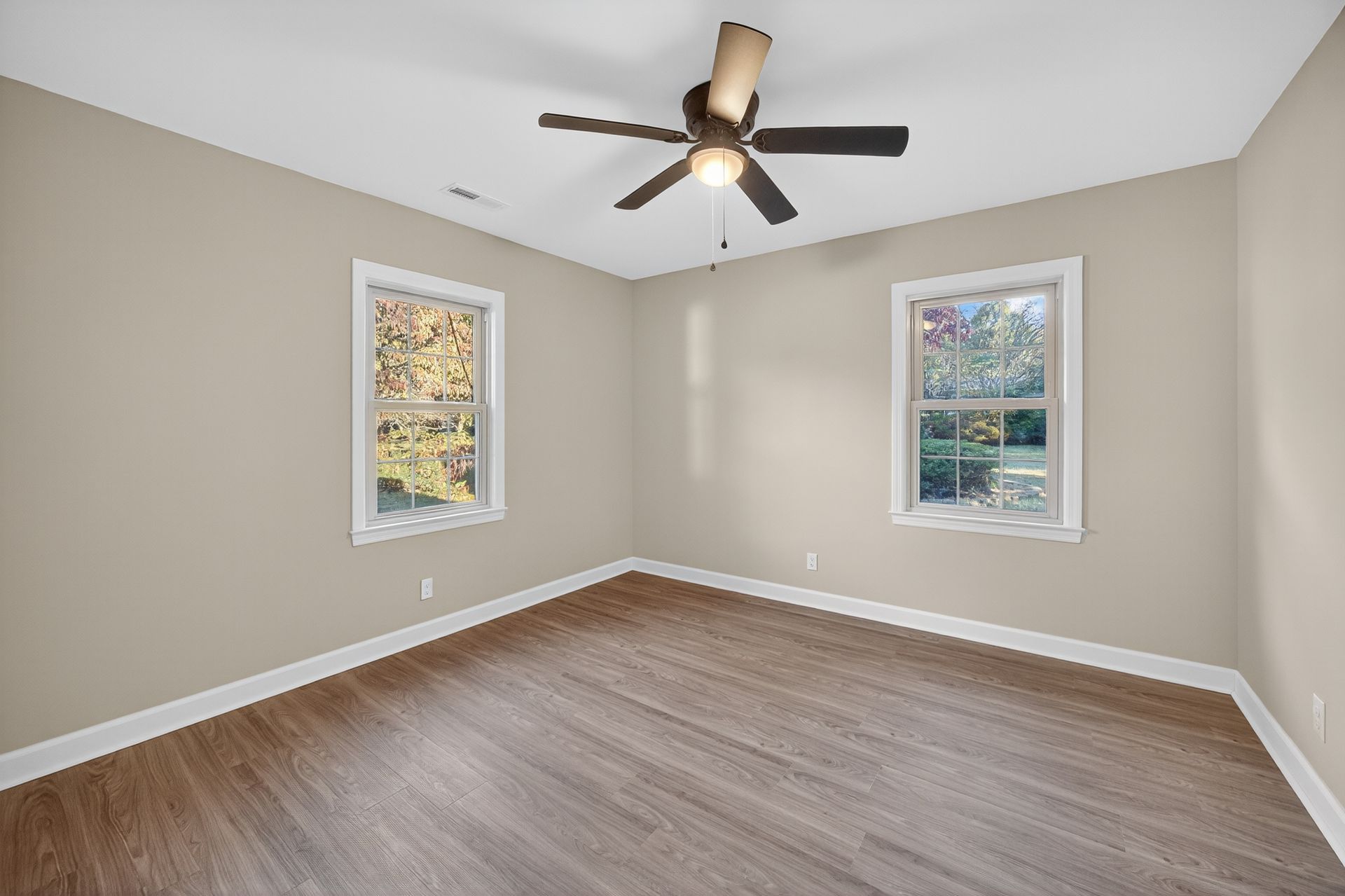 Empty beige bedroom with wood floor, two windows, and a ceiling fan with lights.