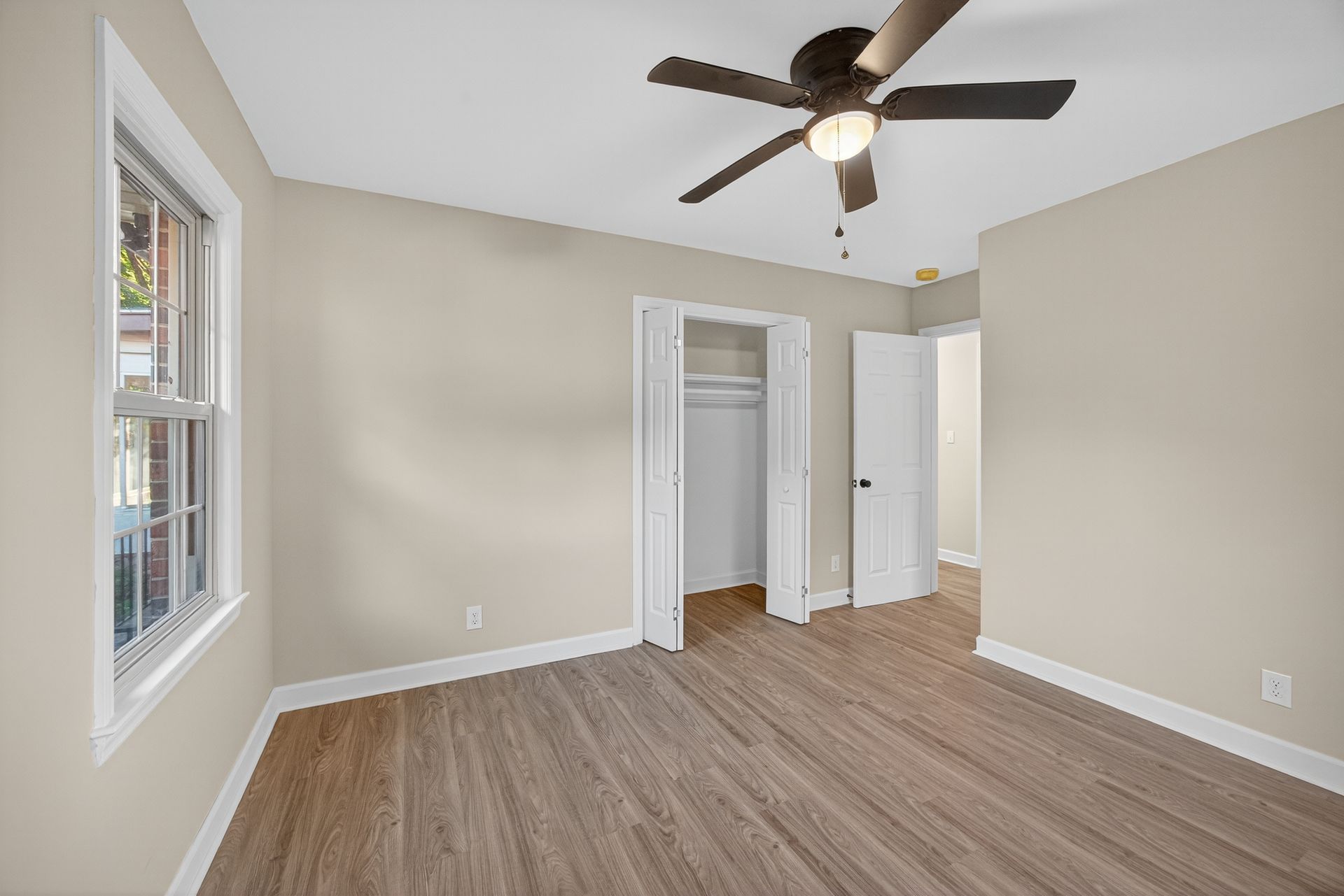 Empty beige bedroom with wood-look flooring, ceiling fan, window, and open closet doors.