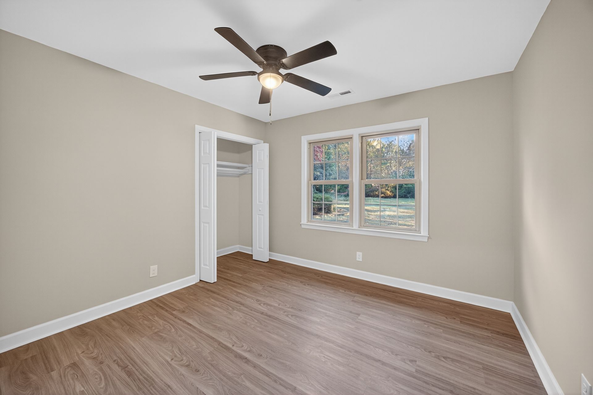 Empty beige bedroom with wood floors, ceiling fan, closet, and a window with white trim