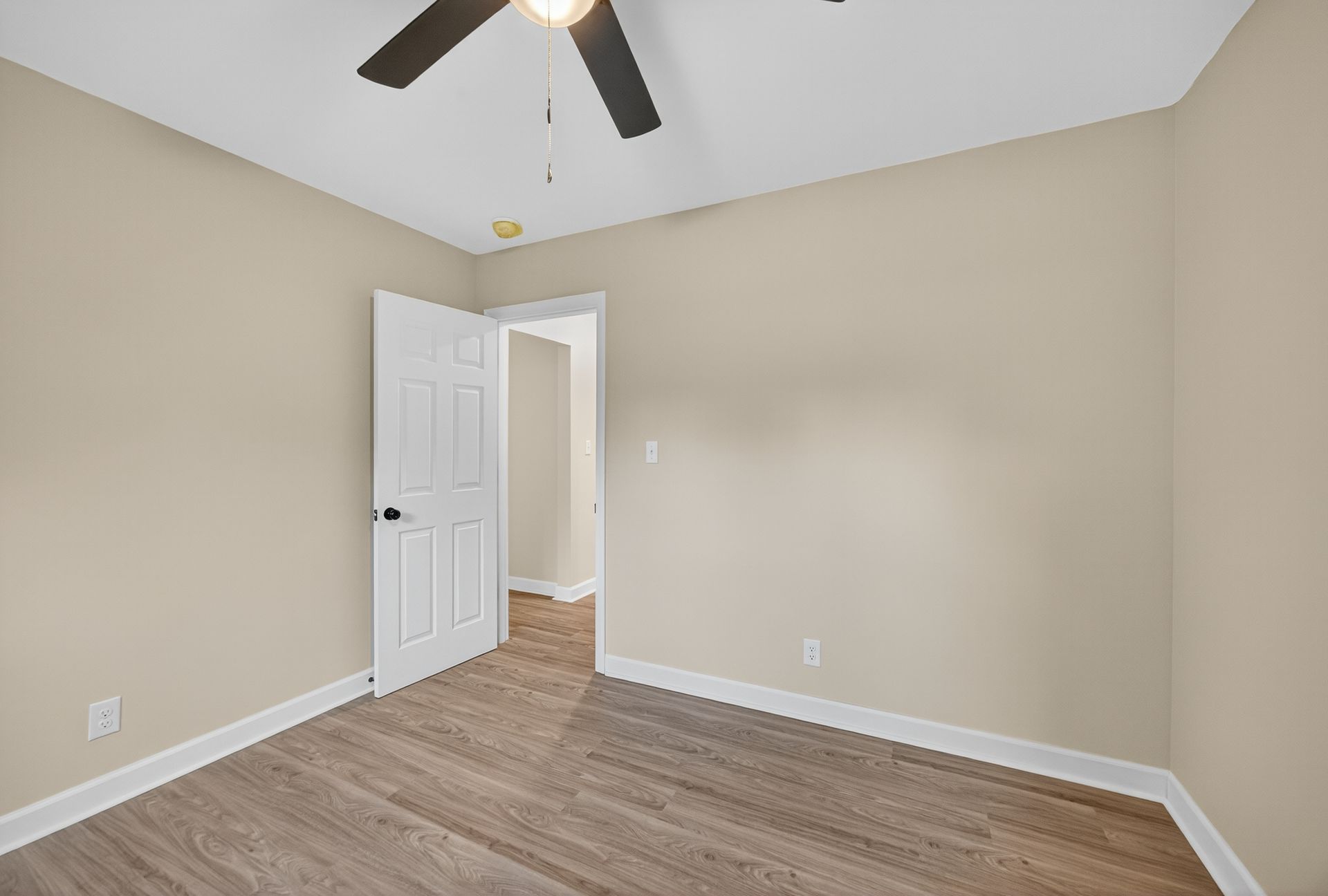 Empty beige bedroom with wood-look flooring, white door, and ceiling fan.