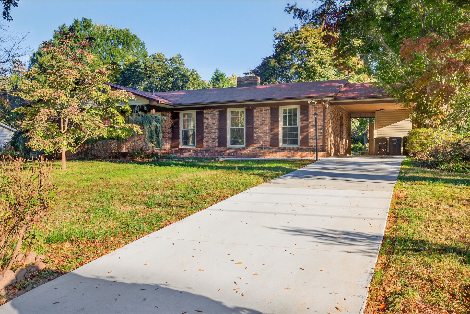Single-story brick house with a concrete walkway and grassy front yard, surrounded by trees.