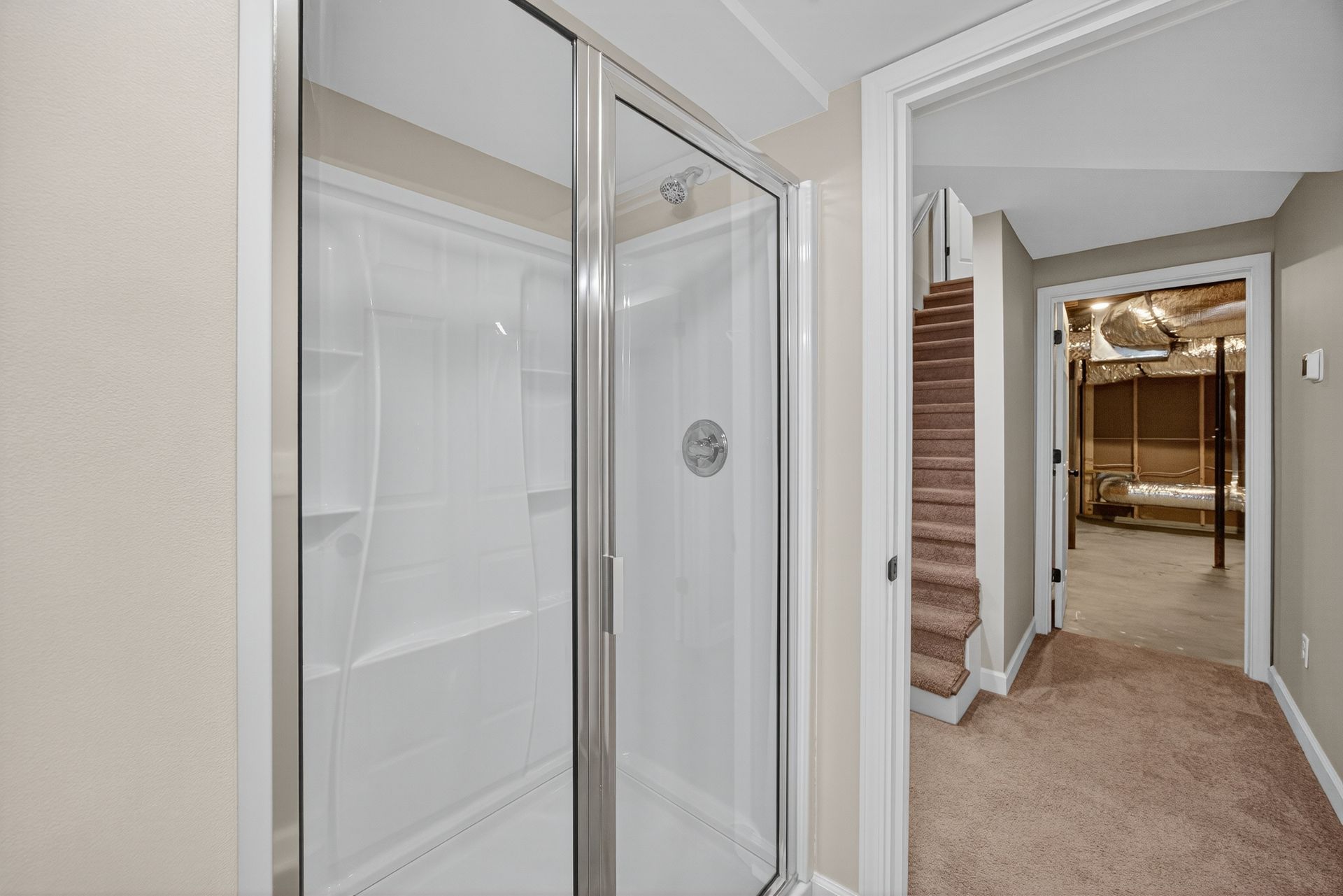 Bathroom with glass shower doors beside a carpeted hallway leading to a basement stairway