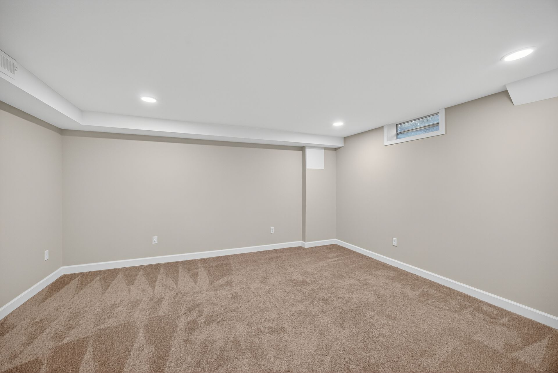 Empty beige carpeted basement room with recessed lights and a small window well.