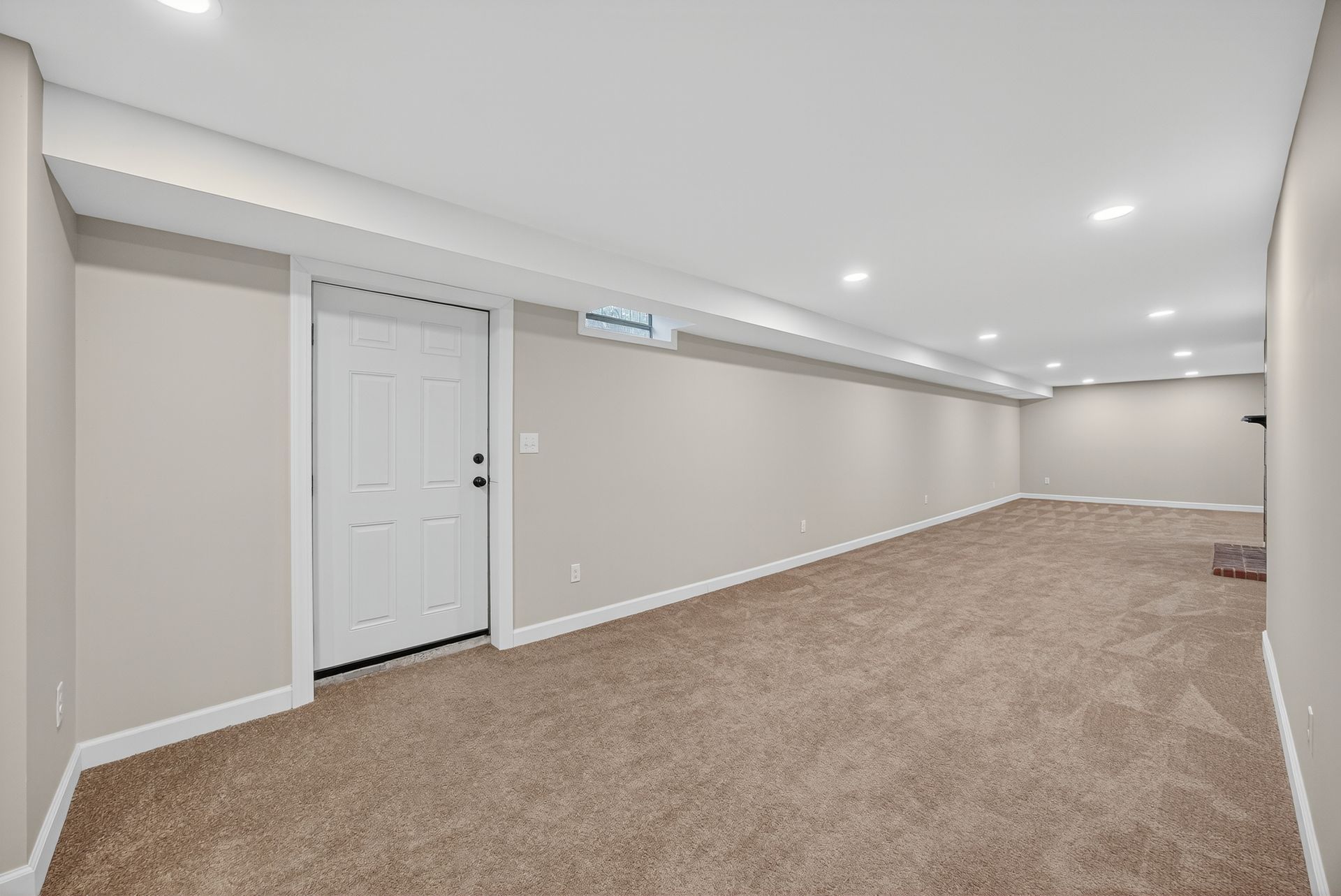 Empty beige carpeted room with white trim, recessed lights, and a white door.
