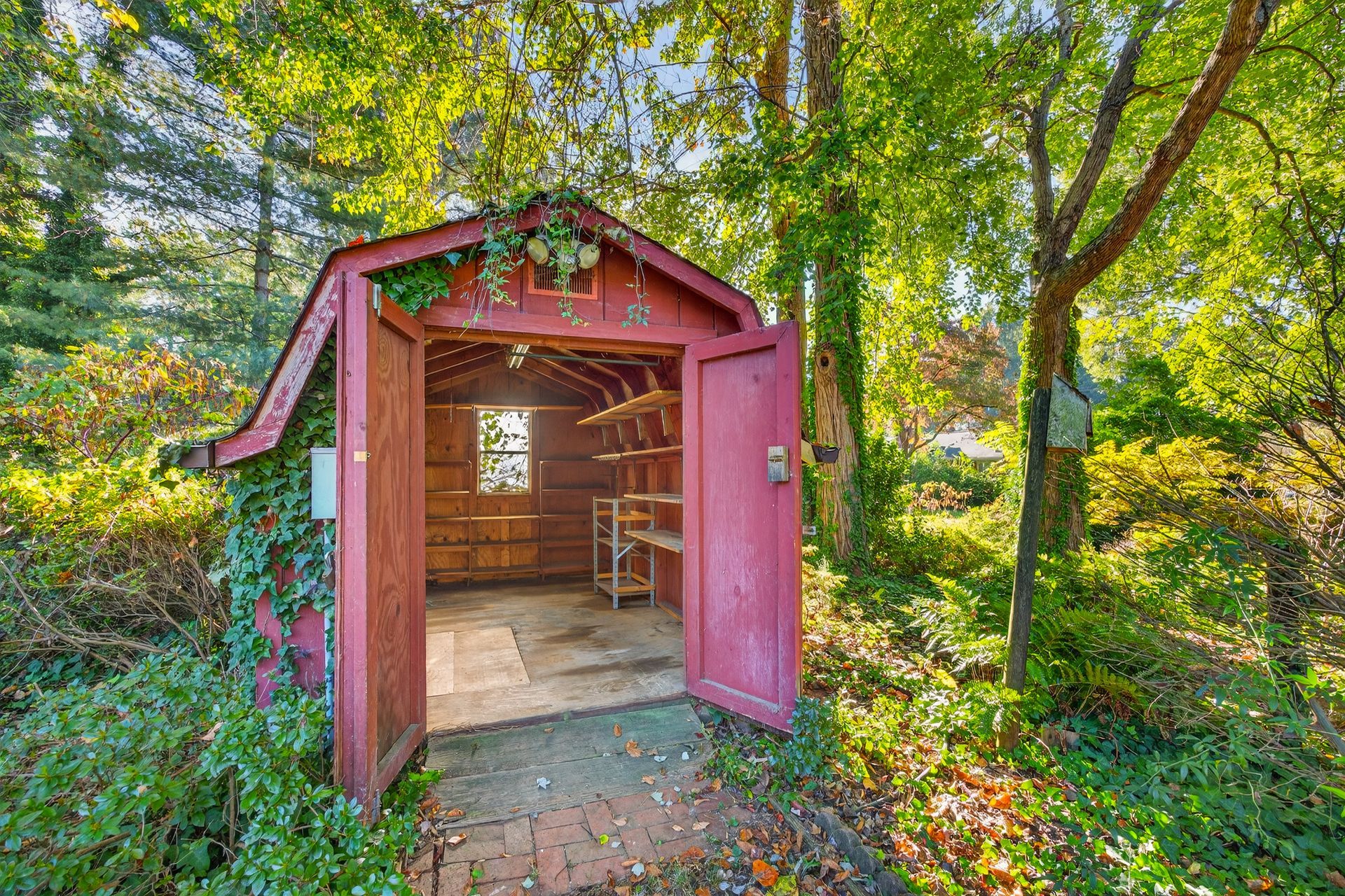 Open red wooden shed in a leafy garden, sunlight streaming through the doorway.