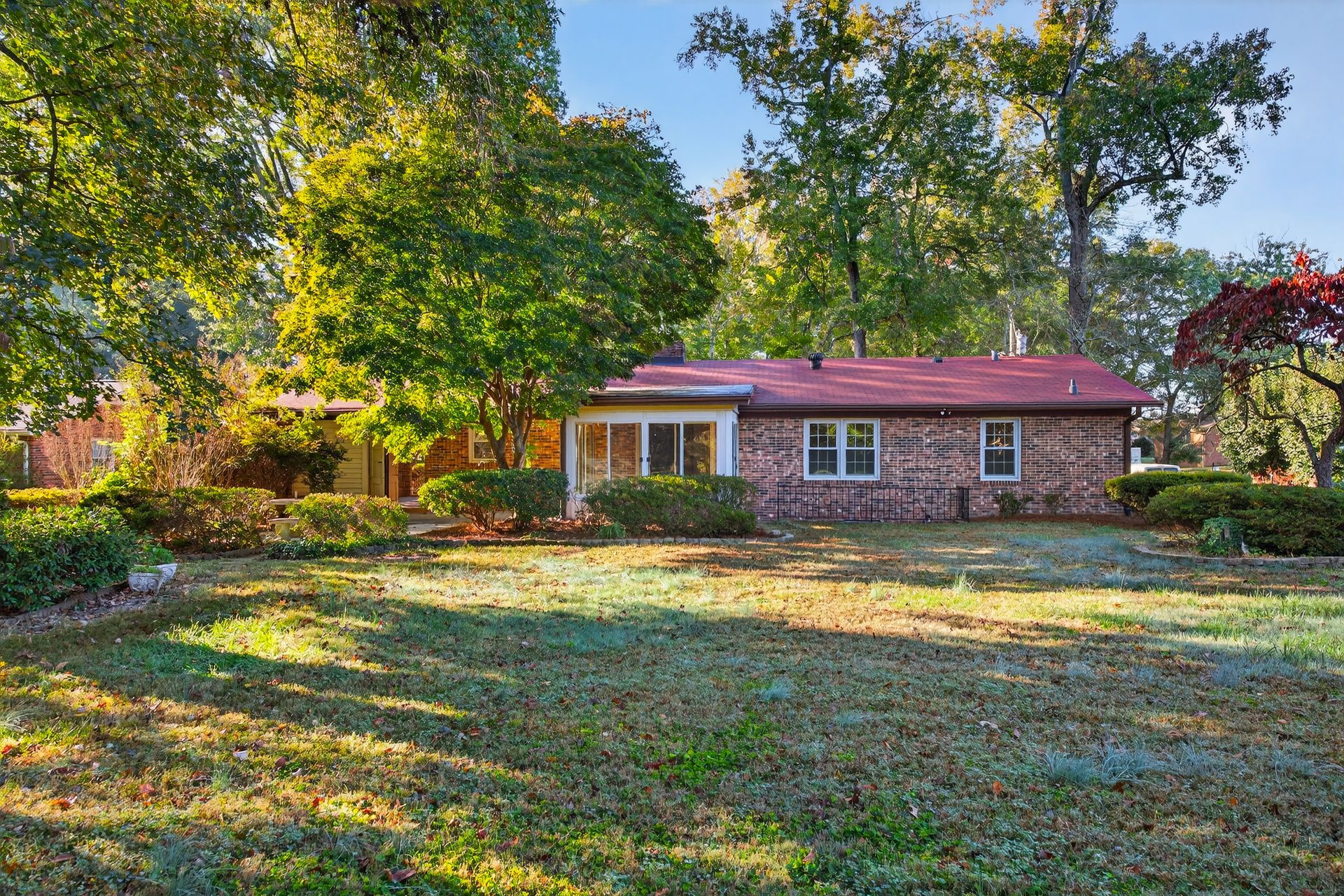 Brick house with red roof in a leafy yard, viewed from a grassy front lawn in autumn sunlight