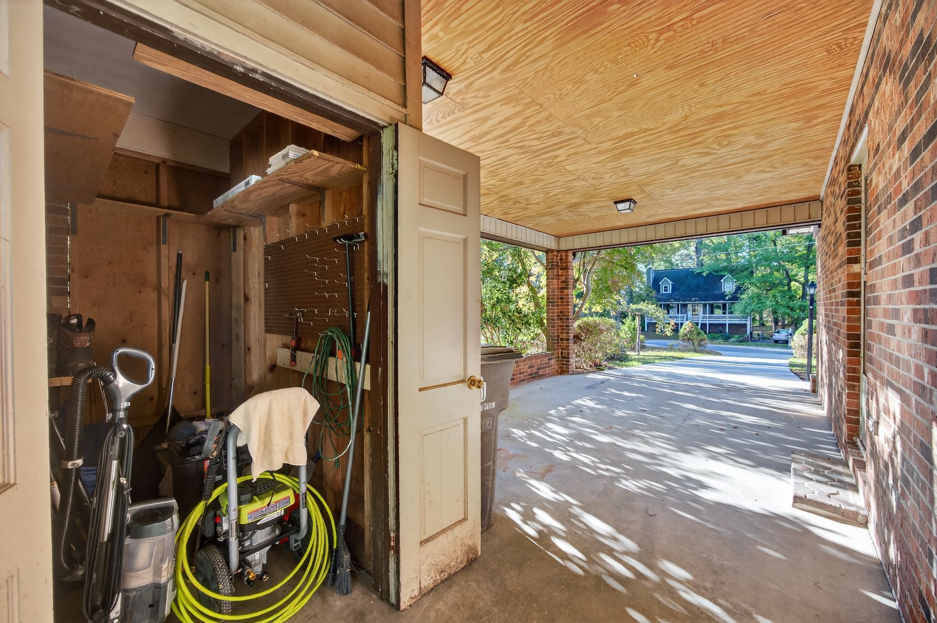 Open garage with tools and hose beside a shaded brick patio overlooking a sunny driveway and blue house.