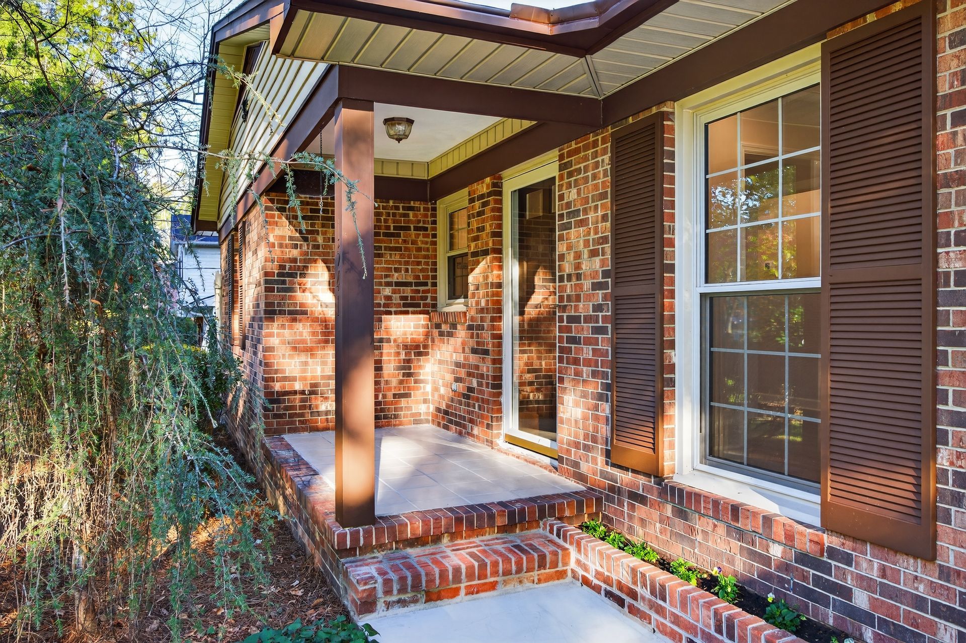 Brick house front porch with white-trimmed windows, wooden shutters, and brick columns in sunlight