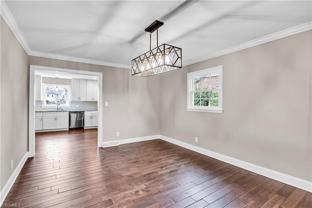 Empty dining room with hardwood floors, beige walls, chandelier, and two windows looking into a kitchen