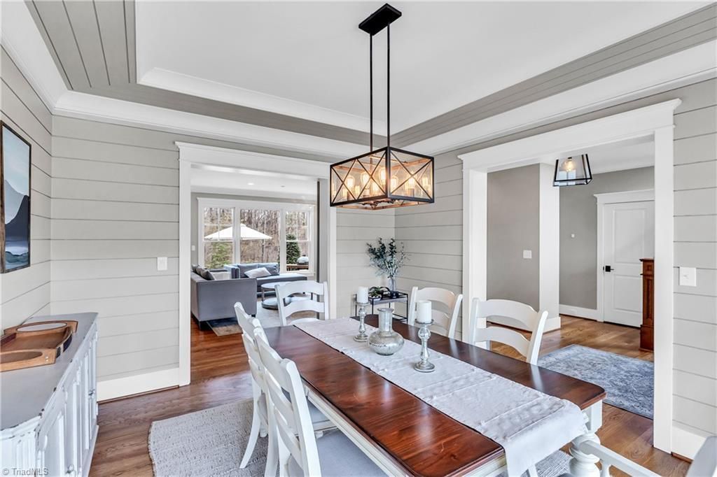 Bright dining room with white chairs, wooden table, and hanging pendant light, opening to a living room and kitchen.