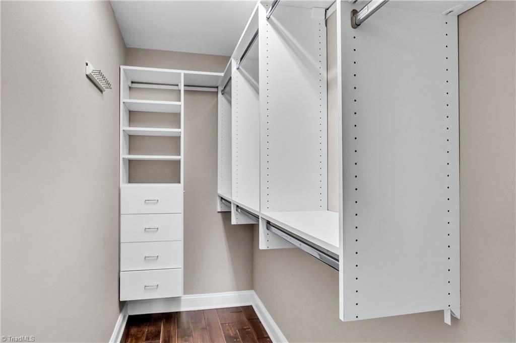 Walk-in closet with white built-in shelves, drawers, and hanging rods on beige walls with dark wood floor
