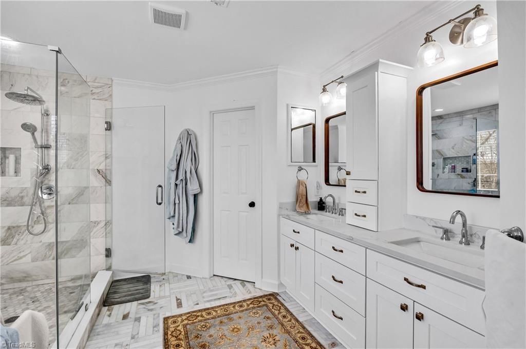 Bright white luxury bathroom with double vanity, large mirrors, glass shower, and striped robe on the door