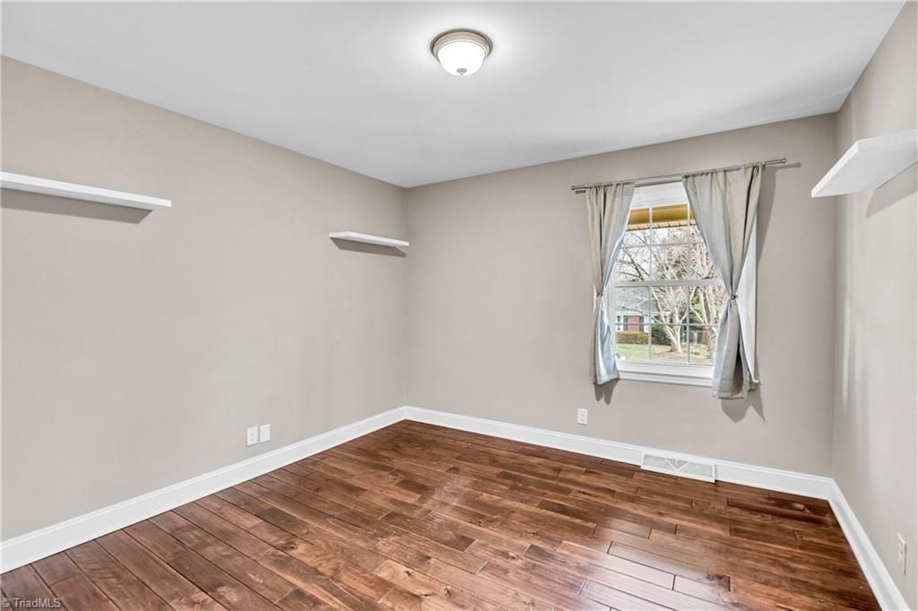 Empty beige bedroom with hardwood floors, a window with curtains, and wall-mounted shelves