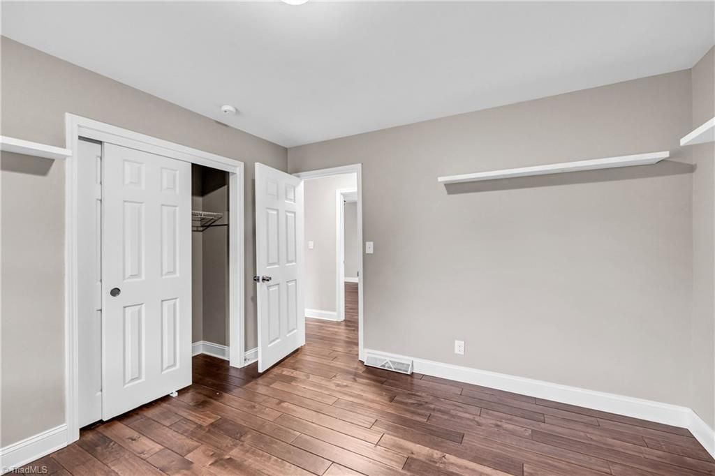 Empty beige bedroom with white closet doors, dark wood floors, and a wall shelf.