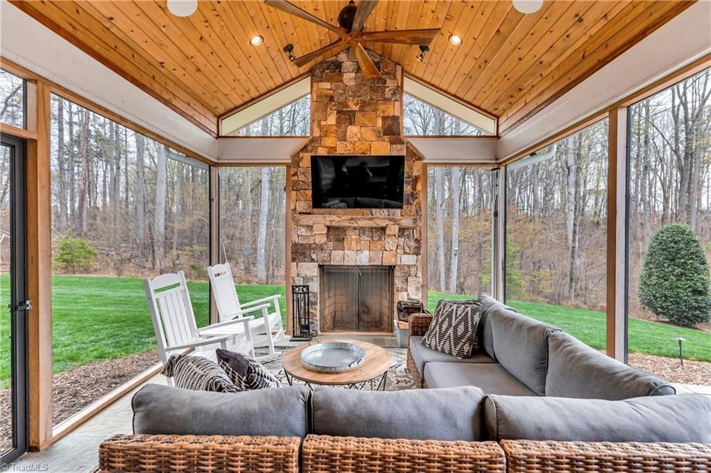 Covered porch with wicker seating around a fire pit and stone fireplace, surrounded by wooded views.