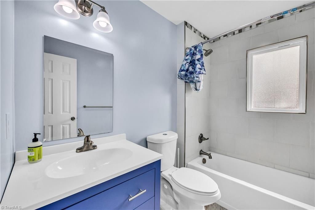 Bright bathroom with blue vanity, white sink and tub, toilet, mirror, and gray walls.
